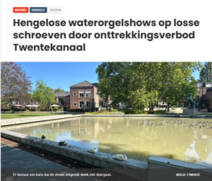 Een stadsvijver met eenden, bomen en flats onder een heldere blauwe lucht, boven een kop over waterorgelshows in Hengelo.